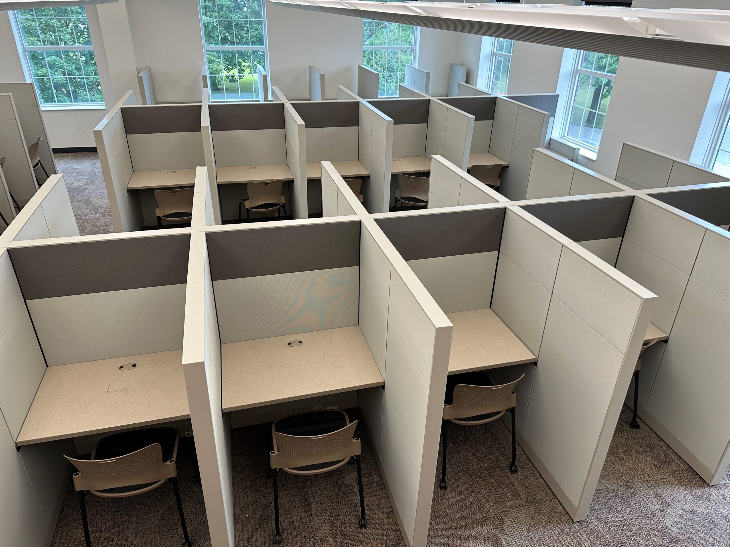 Rows of study cubicles in a bright room, each with a desk and chair. The cubicles are separated by high walls, offering privacy to individual users. Large windows provide views of greenery outside.