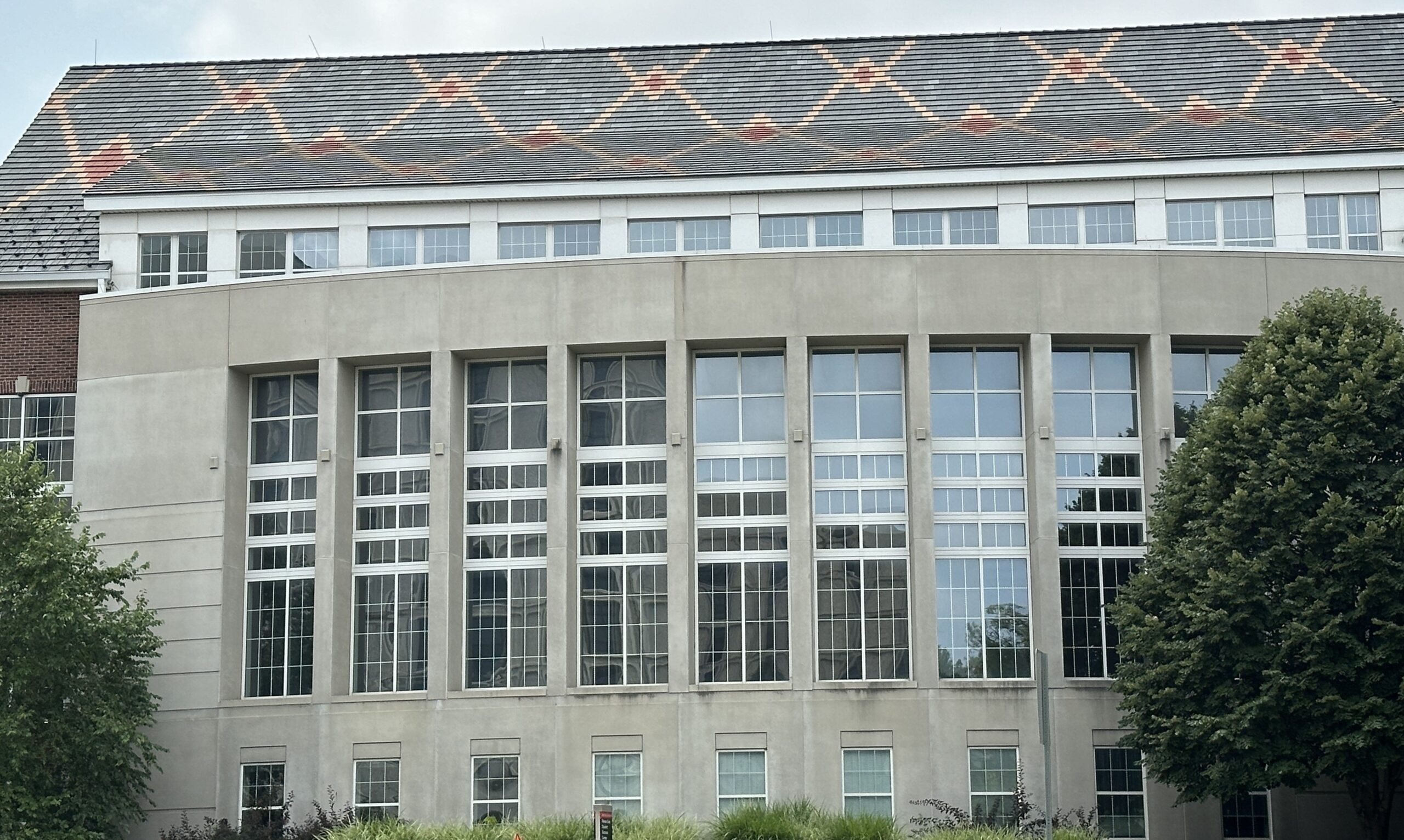 A close-up view of the Hixson-Lied building, highlighting its distinctive roof pattern of red and orange shapes. Below the roof, tall windows line the curved exterior, with greenery such as trees and shrubs completing the scene